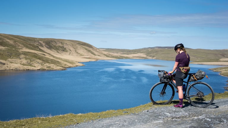Rider pauses beside a moorland lake with Ortlieb handlebar and seat post packs on a gravel bike under a blue sky.