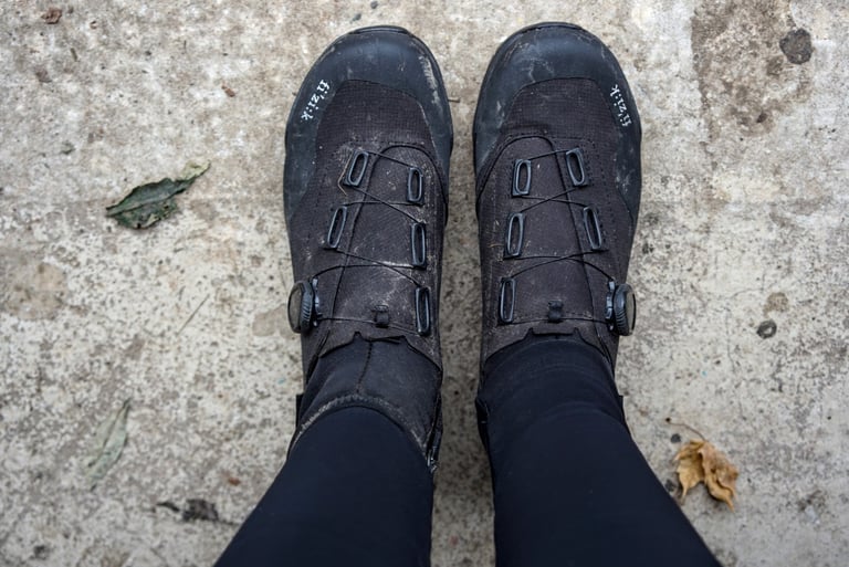 Rider wearing muddy Fizik Terra Artica X2 boots with black tights, seen from above on a concrete path.