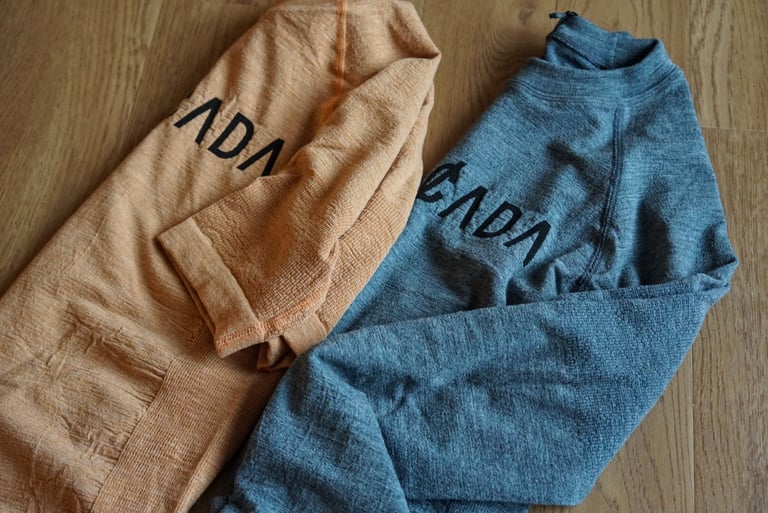 Two folded Cascada merino baselayers, one peach short sleeve and one blue-grey long sleeve, on a wooden floor.