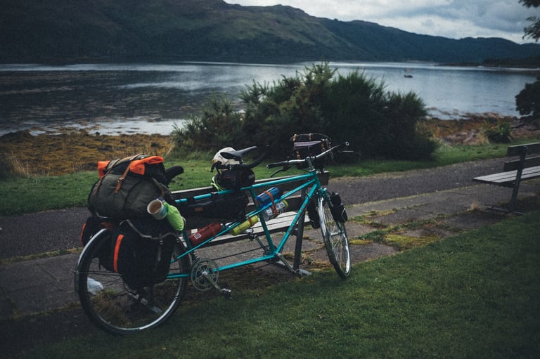 Loaded teal tandem leaning on a bench on the grass beside a sea loch during the resupply stop at Strathcarron.