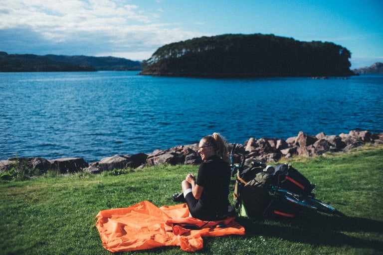 Rider sits on the grass in the sun by the sea at Shieldaig, with an orange poncho and the tandem's panniers alongside.