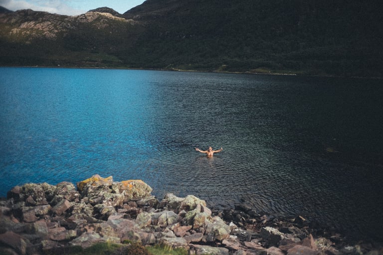 Swimmer with arms outstretched in the blue water of Upper Loch Torridon, with rocks in the foreground.