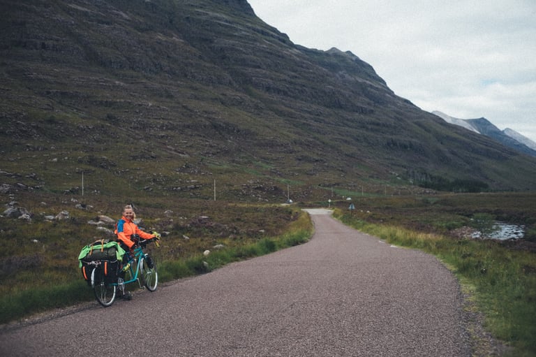 Rider in an orange jacket pauses with the loaded tandem on the low road to Shieldaig below a steep layered hillside.