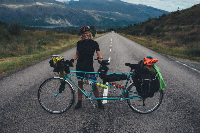 Smiling rider with a helmet and glasses stands behind a loaded teal tandem on the road above Loch Maree.