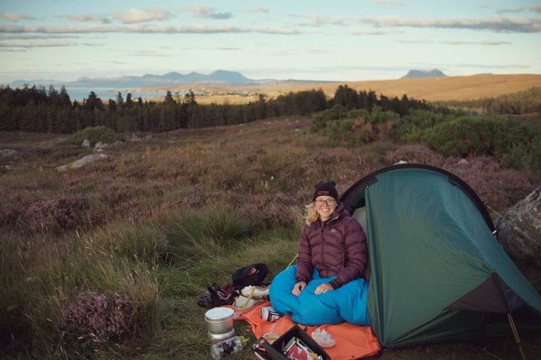 Camper sits in the doorway of the green Zephyros Compact 2 among purple heather, with hills and a coastline behind.