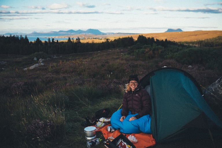 Rider in a beanie sits in a blue sleeping bag at the tent door at a roadside wild camp near Drumchork, coast behind.