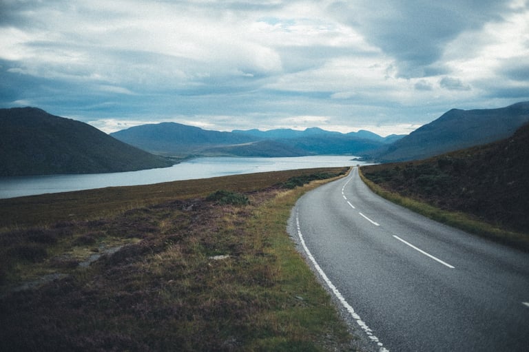 Empty road sweeping downhill along the shore of Little Loch Broom towards mountains under grey cloud.