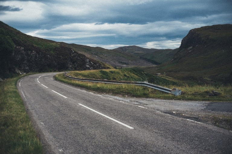 Road bending through a rocky glen with a crash barrier under heavy cloud, on the NC500 near An Teallach.