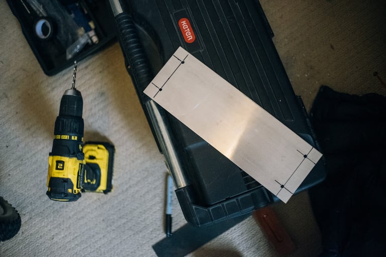 Aluminium plate marked out for drilling, lying on a toolbox beside a cordless drill.