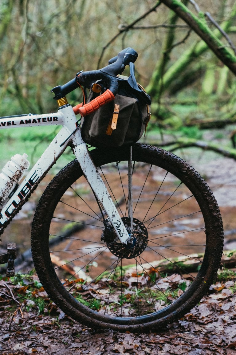Muddy white gravel bike with the Carradice bag on its DIY front support, parked on a wet woodland track.