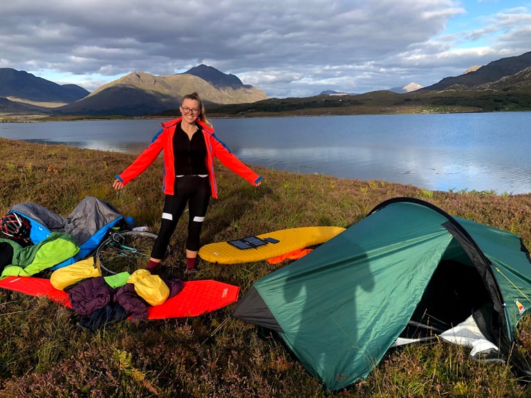 Smiling cyclist by a Zephyros Compact 2 pitched beside a Highland loch, sleeping mats and kit laid out on the heather.