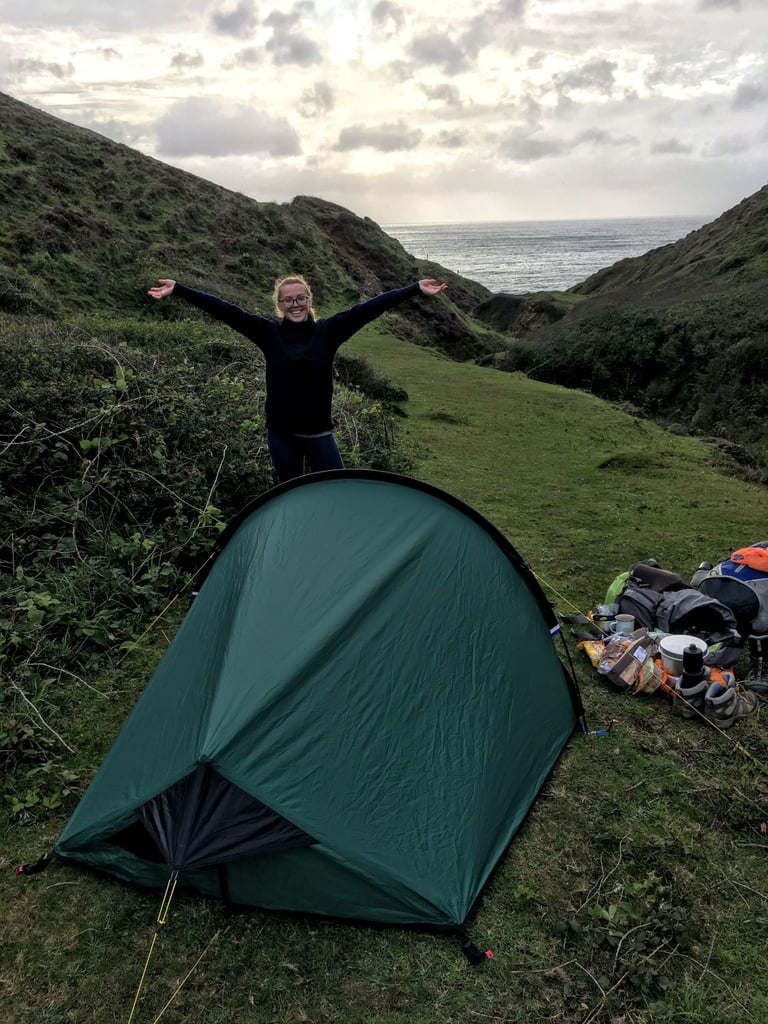 Camper with arms outstretched behind a pitched Zephyros Compact 2 in a Cornish valley that opens onto the sea.