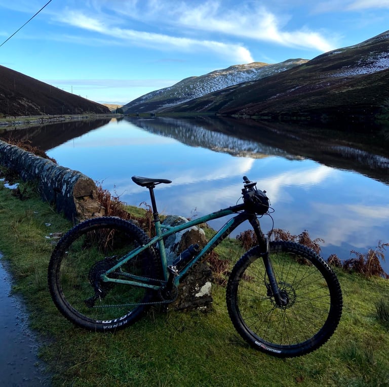 Bombtrack Cale AL leans on a stone wall beside a still reservoir, snow-dusted Pentland Hills reflected in the water.