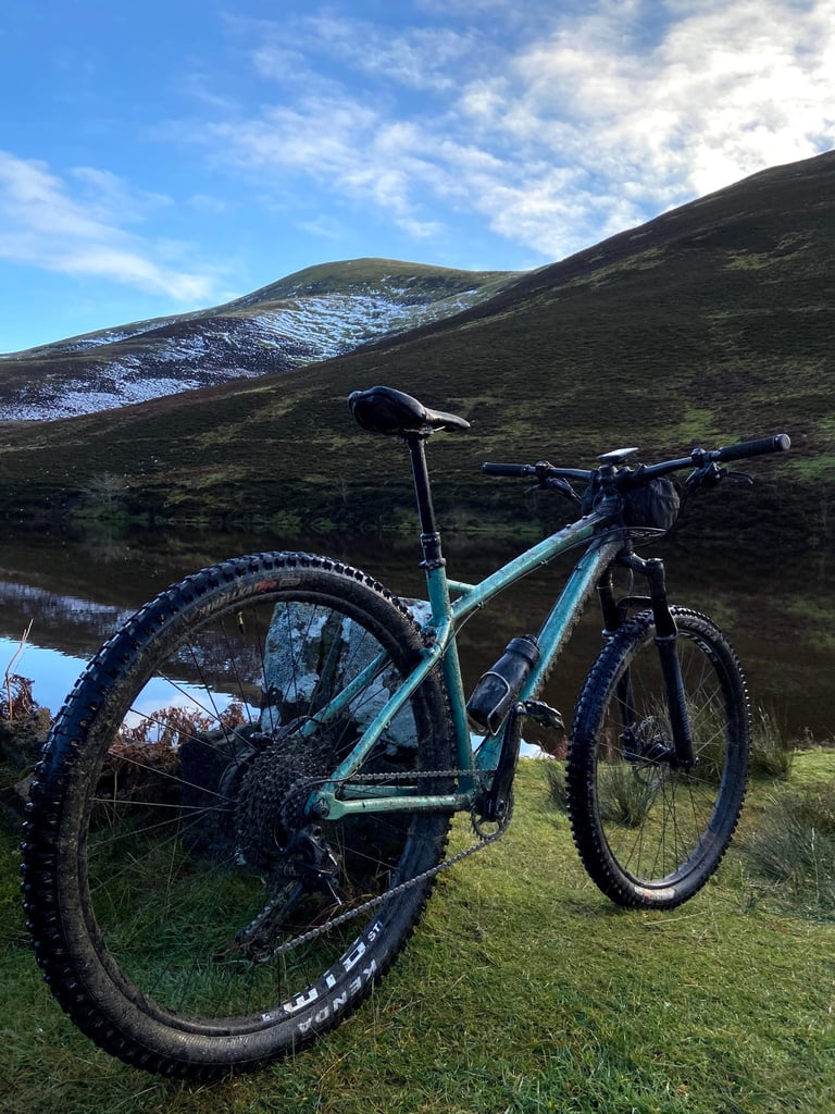 Mud-covered Cale AL propped against rocks on the grassy bank of a Pentland Hills reservoir, where the bike was tested.