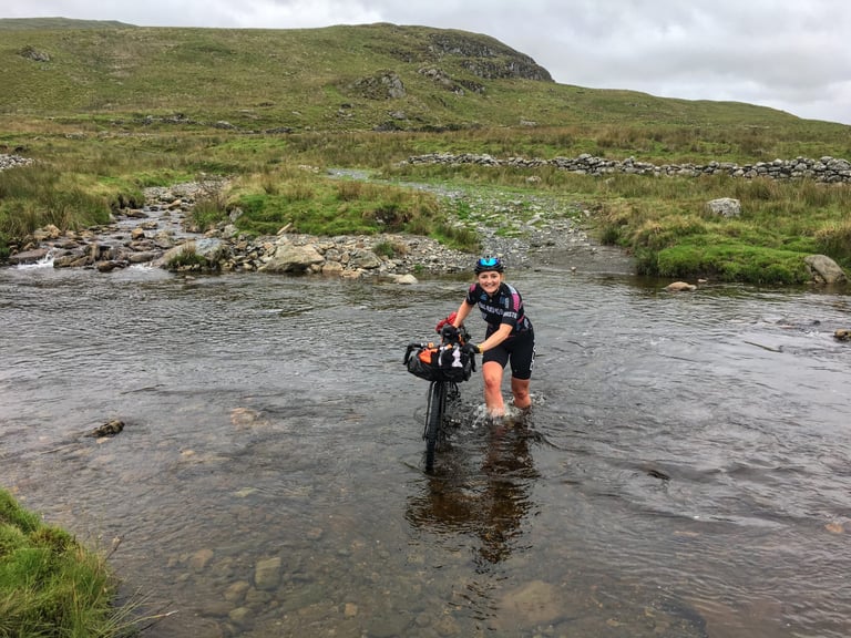 Rider wades a stream pushing a bike with an Ortlieb handlebar pack, with stone walls and moorland behind.