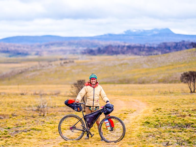 Bikepacker stands with a loaded bike on open grassland in the Jagungal Wilderness, snow-capped peak behind.