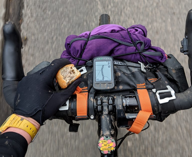Ortlieb handlebar pack with a purple layer held under the front bungee cord, and a sausage roll in a gloved hand.