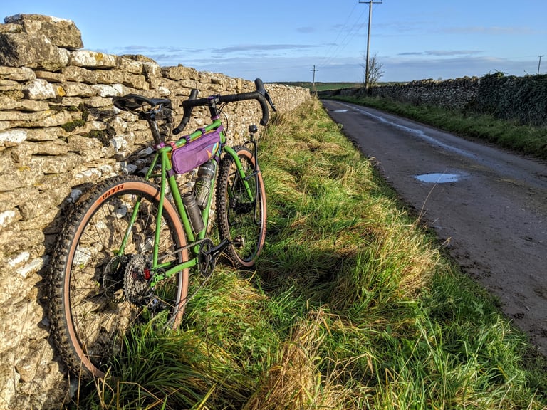 Green Fairlight Faran with a purple half frame bag leaning on a Cotswold stone wall beside a muddy lane.