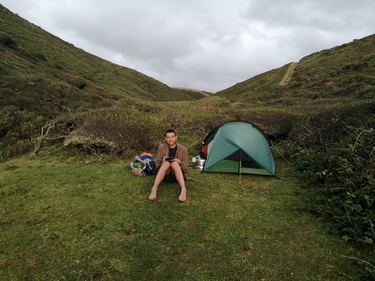 Barefoot camper sits beside a pitched Zephyros Compact 2 in a grassy valley under grey cloud, with kit and pots nearby.