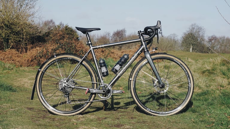 Planet X Tempest set up for road riding with 700c wheels, tan-wall tyres and mudguards, on grass beside bracken.
