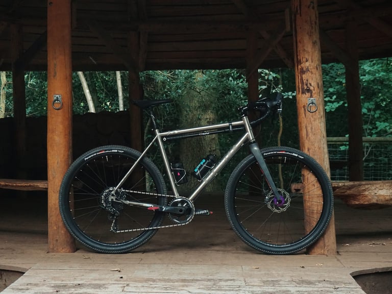 Planet X Tempest Ti gravel bike with flared bars and purple Hope hub, standing in a timber-framed shelter.