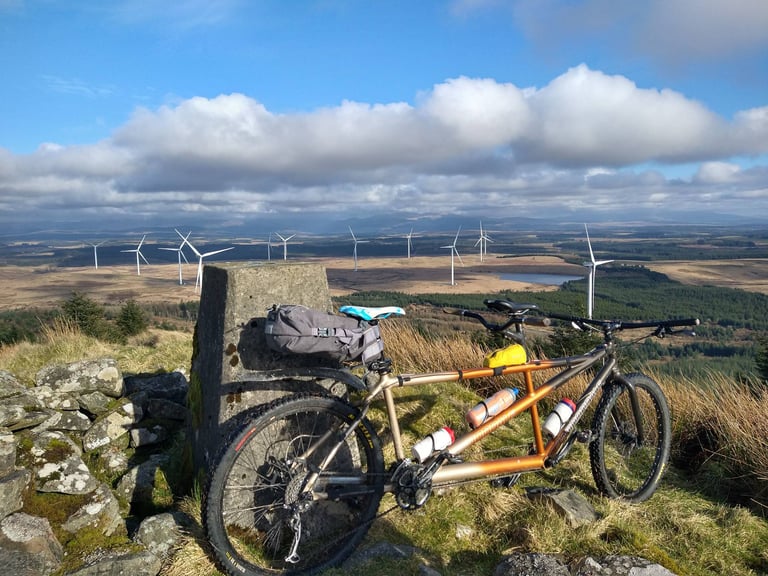 Tandem mountain bike leaning on a trig pillar on a hilltop above forest and a wind farm, one of three tandem entries.