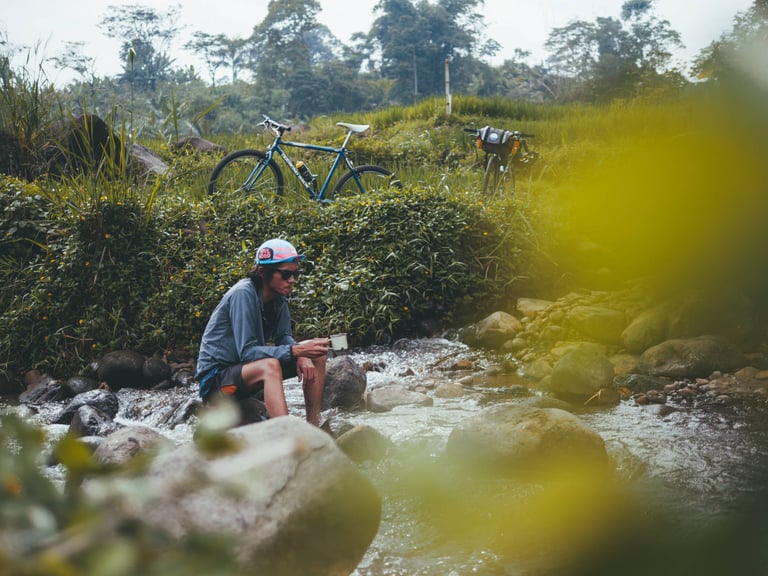 Rider sits on rocks beside a stream with a mug, two bikes parked in green fields behind, near Bogor, Indonesia.