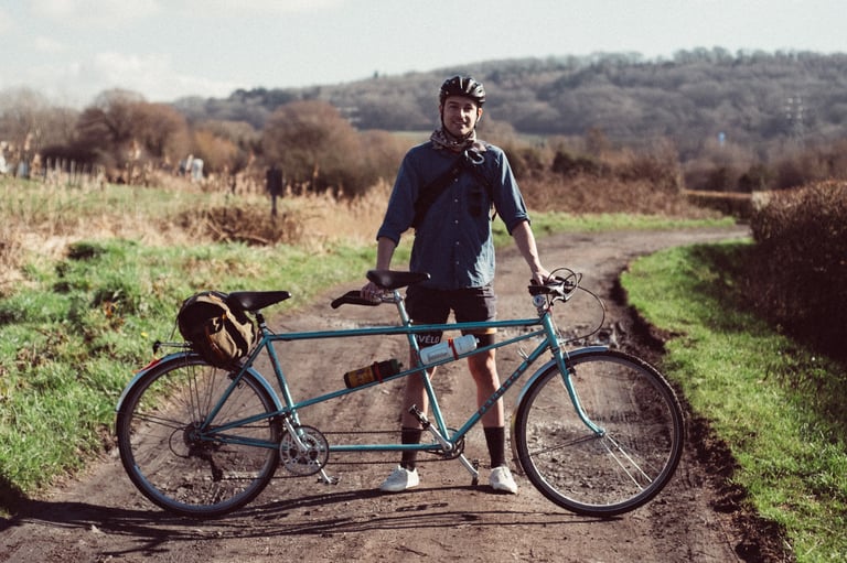Rider in a helmet stands behind a teal steel tandem on a dirt lane, the bike that now runs the SimWorks Bubbly pedals.