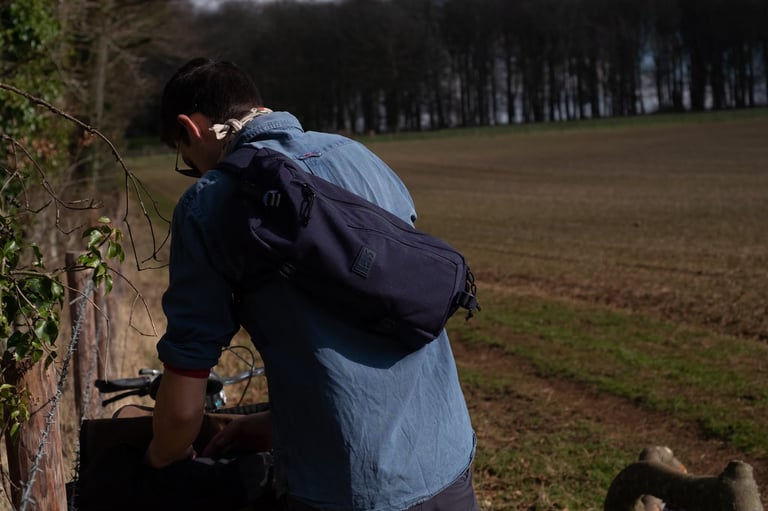 Chrome Kadet sling bag in Blue Tonal on a rider's back as they lean over a bike beside a bare field and fence.