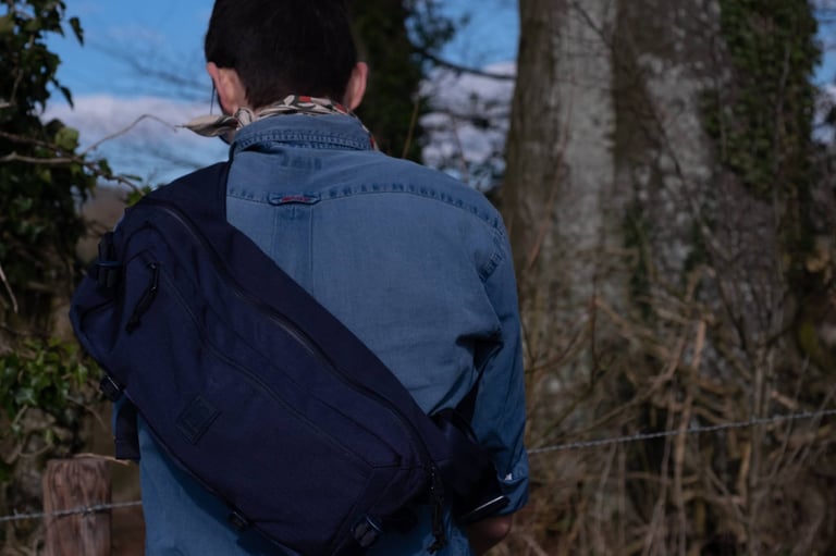 Navy Chrome Kadet sling bag worn across the back of a denim shirt, seen from behind beside a wire fence.