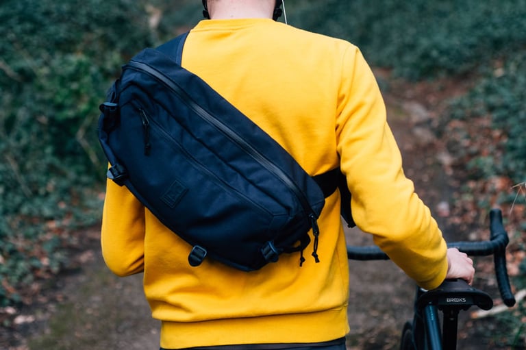 Chrome Kadet sling bag worn over a yellow sweatshirt, rear view, as the wearer wheels a drop-bar bike down a leafy path.