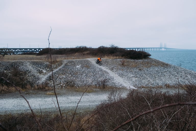 Rider in an orange jacket on a gravel track by the sea with a long bridge behind, on a wintery Swedish ride for the event.
