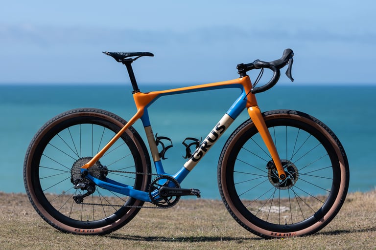 Finished GRUS bamboo gravel bike in orange, blue and cream with a Shimano GRX drivetrain and Praxis crank, by the sea.