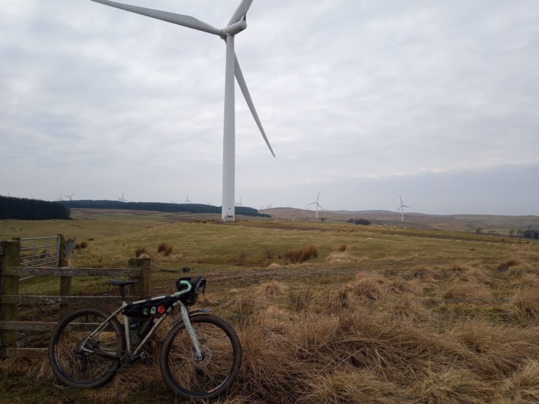 Gravel bike with a frame bag leans on a wooden fence beneath a wind turbine on the Glasgow Inner Circle route.