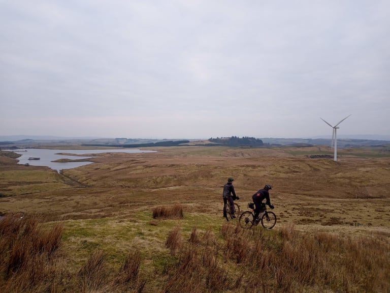 Two riders cross open moorland past a reservoir and wind turbine on the Glasgow Inner Circle route.