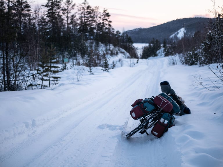 Loaded touring bike lies in deep snow beside a snowy road near Lake Baikal, Siberia, panniers and boots visible.