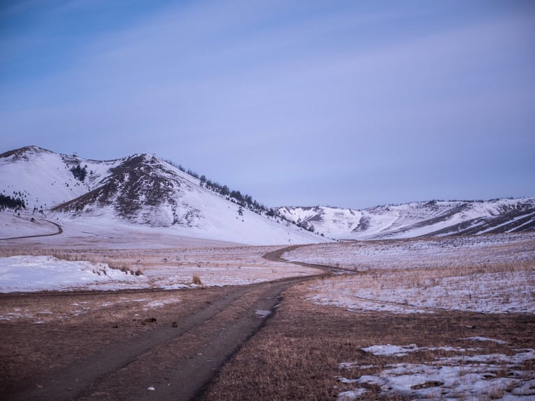 Empty dirt track crosses snow-patched hills under a pale blue sky in winter, Lake Baikal region.