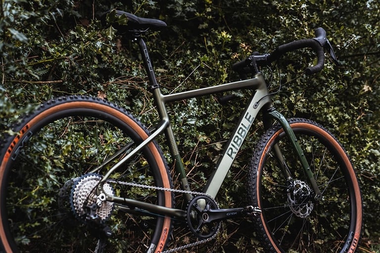 Ribble Gravel AL leaning in a holly hedge, showing its olive-green alloy frame, GRX cranks and tan-wall tyres.