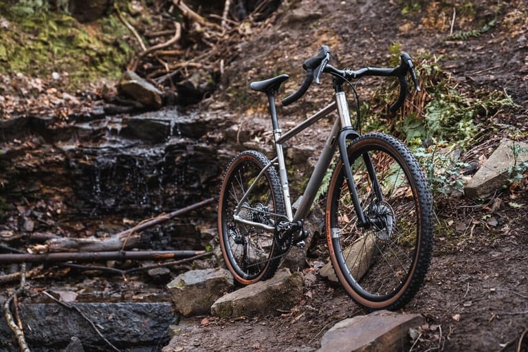 Ribble Gravel Ti with a brushed titanium frame parked on a rocky woodland trail beside a small waterfall.