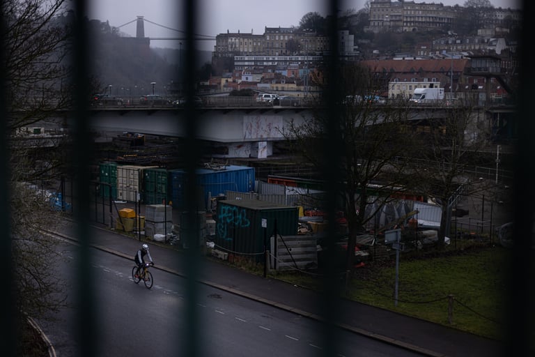 Lone cyclist on a wet Bristol road below the Clifton Suspension Bridge, seen through railings, in The Tipping Point.