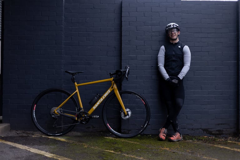 Rider Fergus Ryan stands beside a yellow Santa Cruz gravel bike against a dark brick wall.