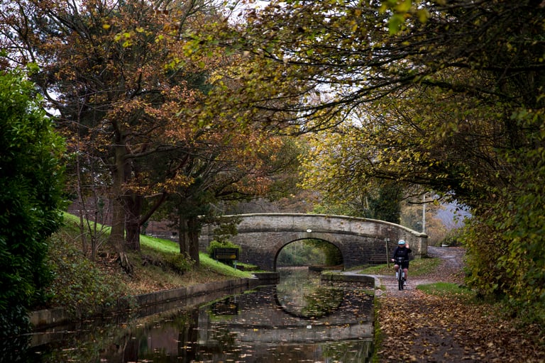Cyclist on a canal towpath passes a stone arched bridge beneath autumn trees near Llangollen, North Wales.