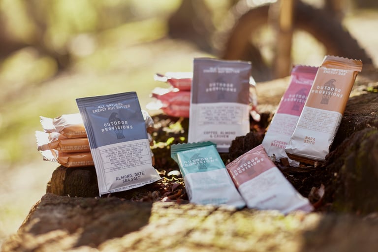 Outdoor Provisions nut butter sachets and four flavours of energy bar laid out on a tree stump.