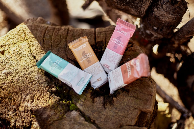 Outdoor Provisions energy bars in four flavours, Choc Kendal Mint Cake, Bara Brith, Cherry Bakewell and Parkin, on a stump.