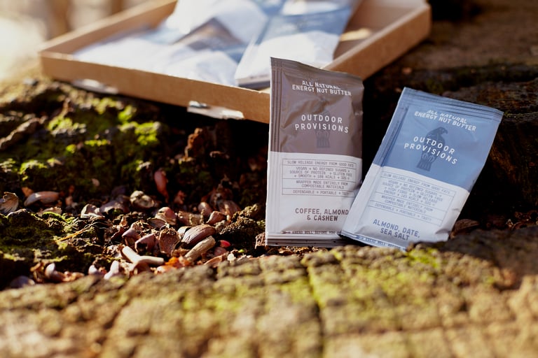 The two Outdoor Provisions nut butter flavours stood on a mossy log, their cardboard delivery box behind.