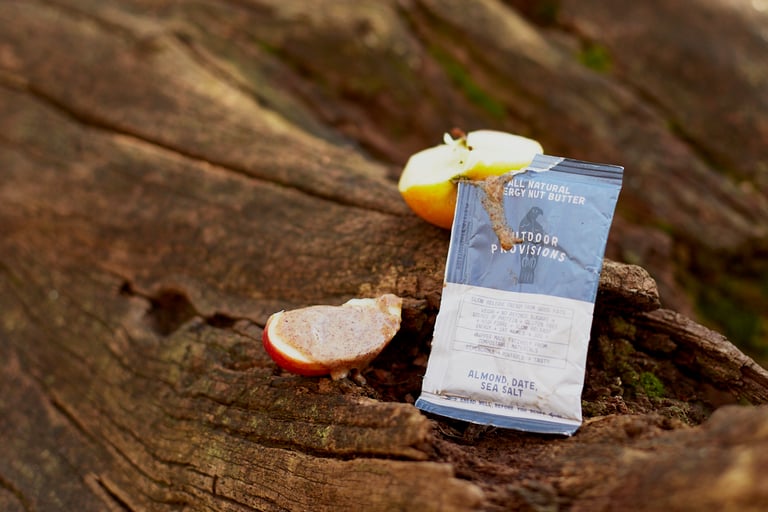 Opened Outdoor Provisions nut butter sachet propped on a log next to apple slices spread with almond butter.