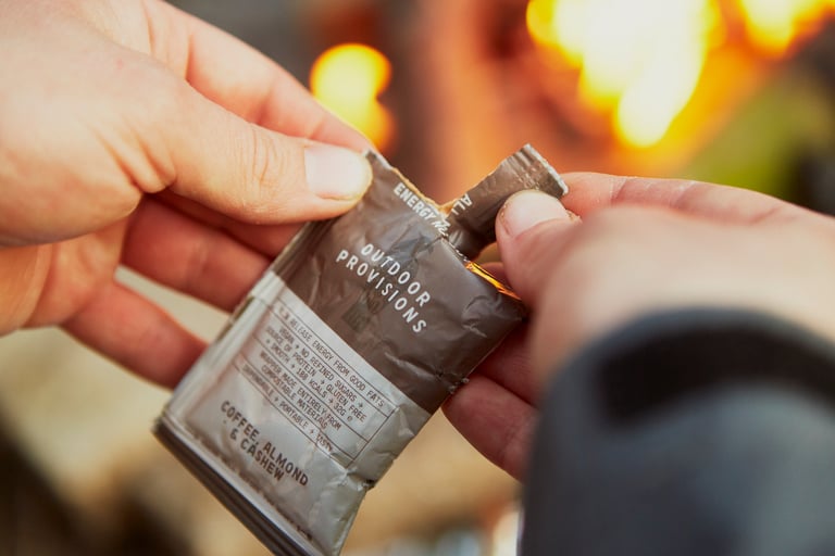 Hands tearing open a coffee, almond and cashew Outdoor Provisions nut butter sachet in front of a glowing fire.