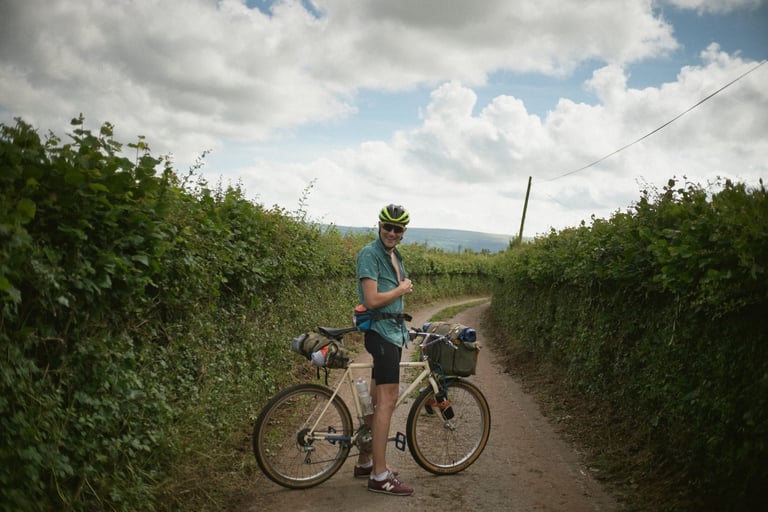 Rider stands with a bag-laden cream Specialized Rockhopper on a narrow hedge-lined lane under a cloudy sky.