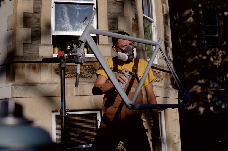 Rider in a respirator holds a spray can beside a grey primer-coated frame on a stand, giving an old MTB a home repaint.