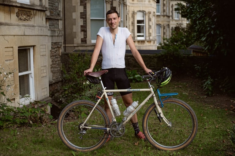 Smiling rider stands beside a cream Specialized Rockhopper converted into a gravel bike, with a blue front rack.